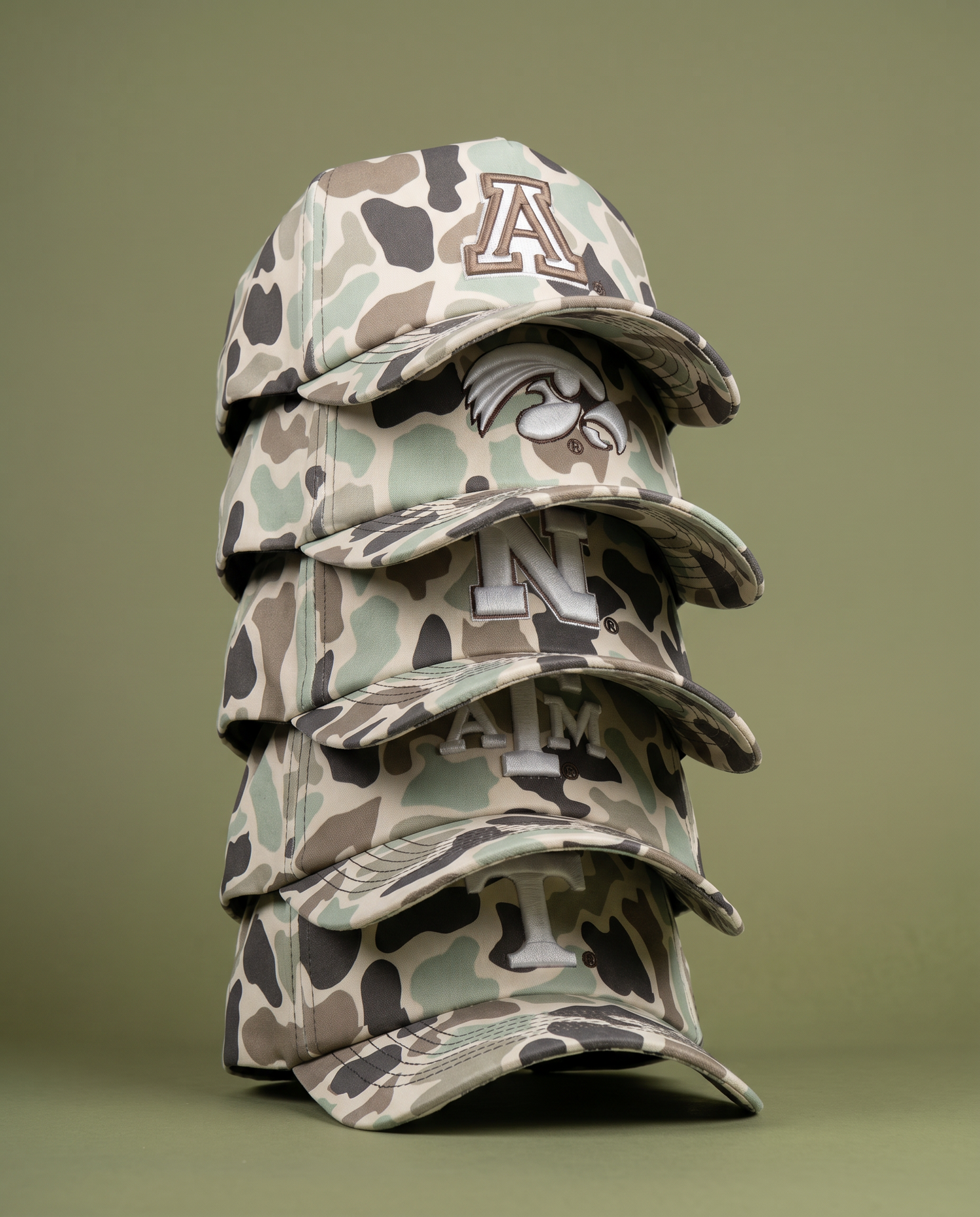 A stack of five camouflage baseball caps with different embroidered college logos on the front, arranged against a plain olive-green background.