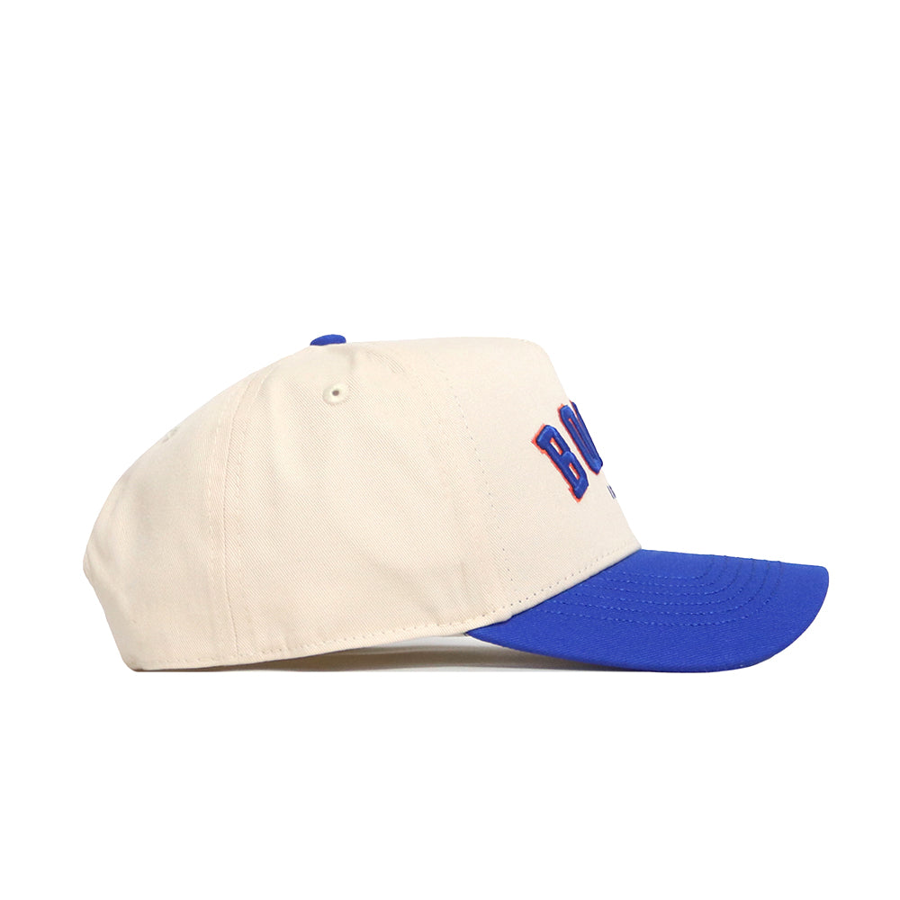The No Rivals Boise Hometown Hat is a cream cotton baseball cap with a blue curved brim and embroidered front letters, pictured from the left side on a white background.