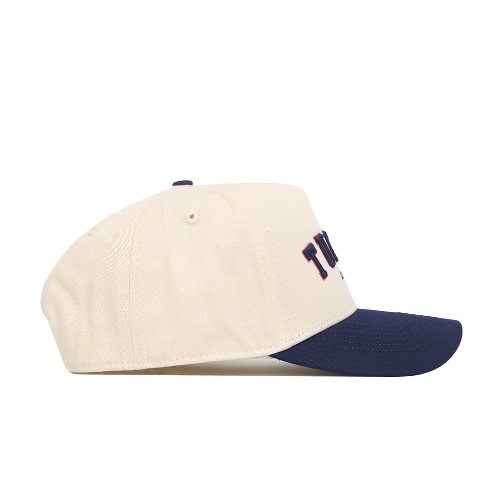 The No Rivals Tucson Hometown Hat is a cream-colored snapback with a navy blue brim, seen from the side. The front has partially visible dark embroidery and a structured fit crafted from soft cotton fabric.