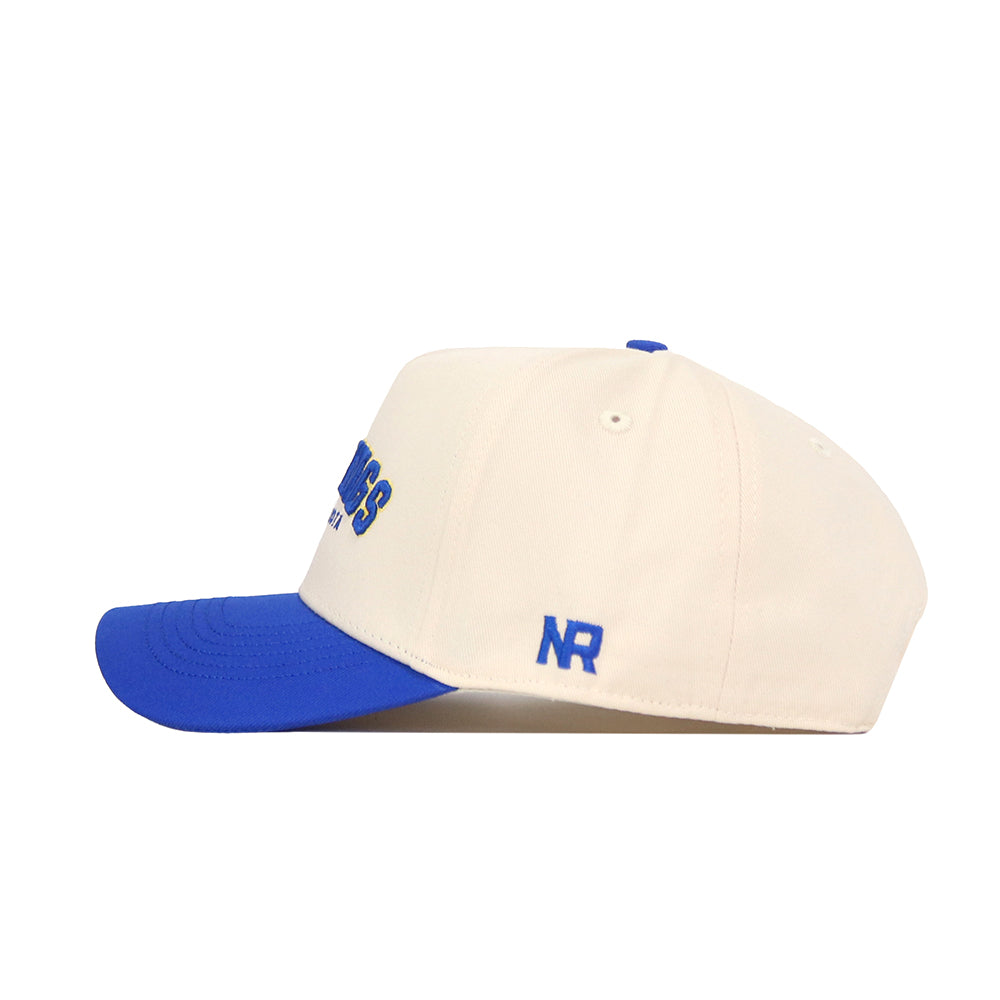 The No Rivals Brookings Hometown Hat is a cream snapback with a blue brim and button, featuring blue NR embroidery on the side and partial blue text on the front. Made from high quality cotton, it is shown here in side profile.