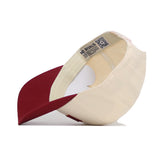 The Ames Hometown Hat by No Rivals is shown upside down, revealing the inside tag and a QR code on the inner band. This cream and burgundy snapback features a structured fit, flat brim, round crown, and high-quality cotton construction.