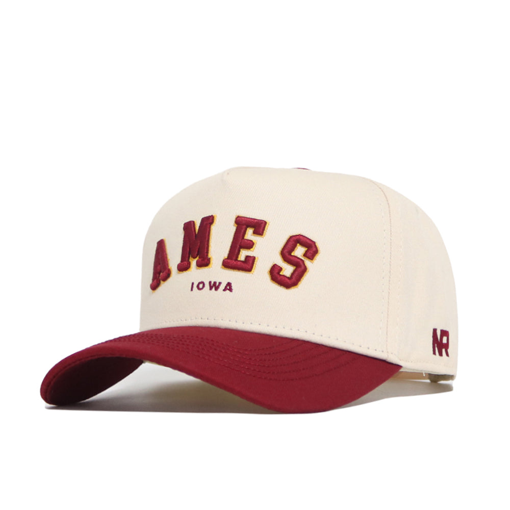The No Rivals Ames Hometown Hat is a cream cotton baseball cap with a maroon brim, featuring bold maroon and yellow "AMES" and "IOWA" on the front, plus a small maroon NR logo on the side.