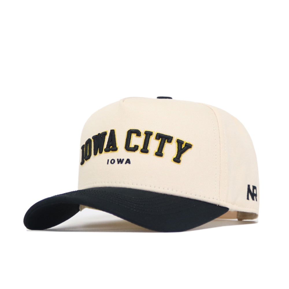 The No Rivals Iowa City Hometown Hat is a structured beige cotton baseball cap with a black brim, bold yellow and black IOWA CITY lettering above IOWA, NR on the side, and an adjustable snapback for comfort.