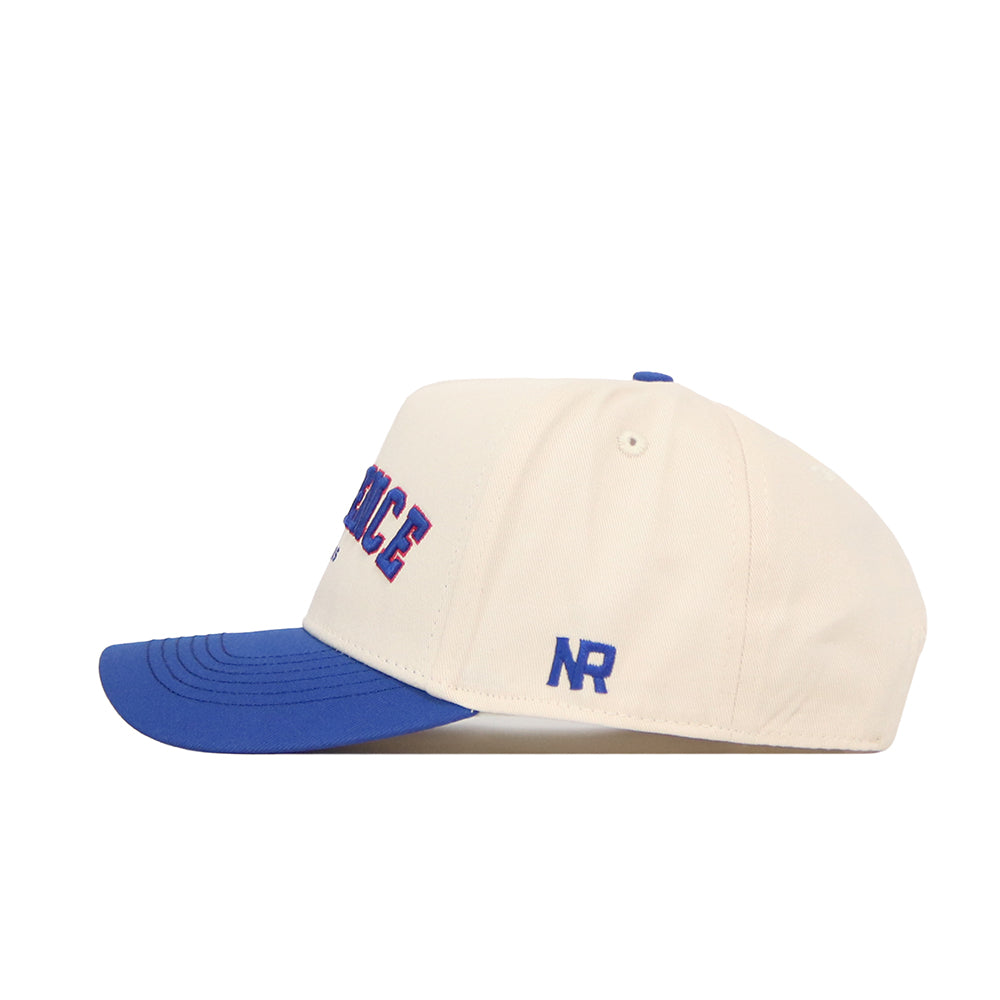 The Lawrence Hometown Hat by No Rivals is a cream snapback with a blue brim, made of high-quality cotton. It features blue and red embroidered text on the front and “NR” stitched in blue on the side for a structured fit.