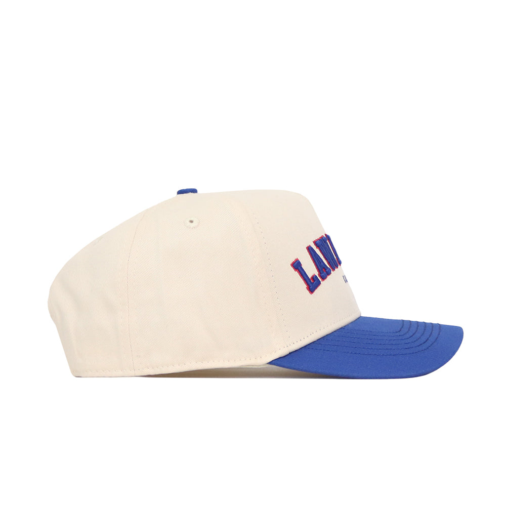 The No Rivals Lawrence Hometown Hat is a cream cotton baseball cap with a blue curved brim and "LAND" embroidered in blue and red, shown side profile on a white background.