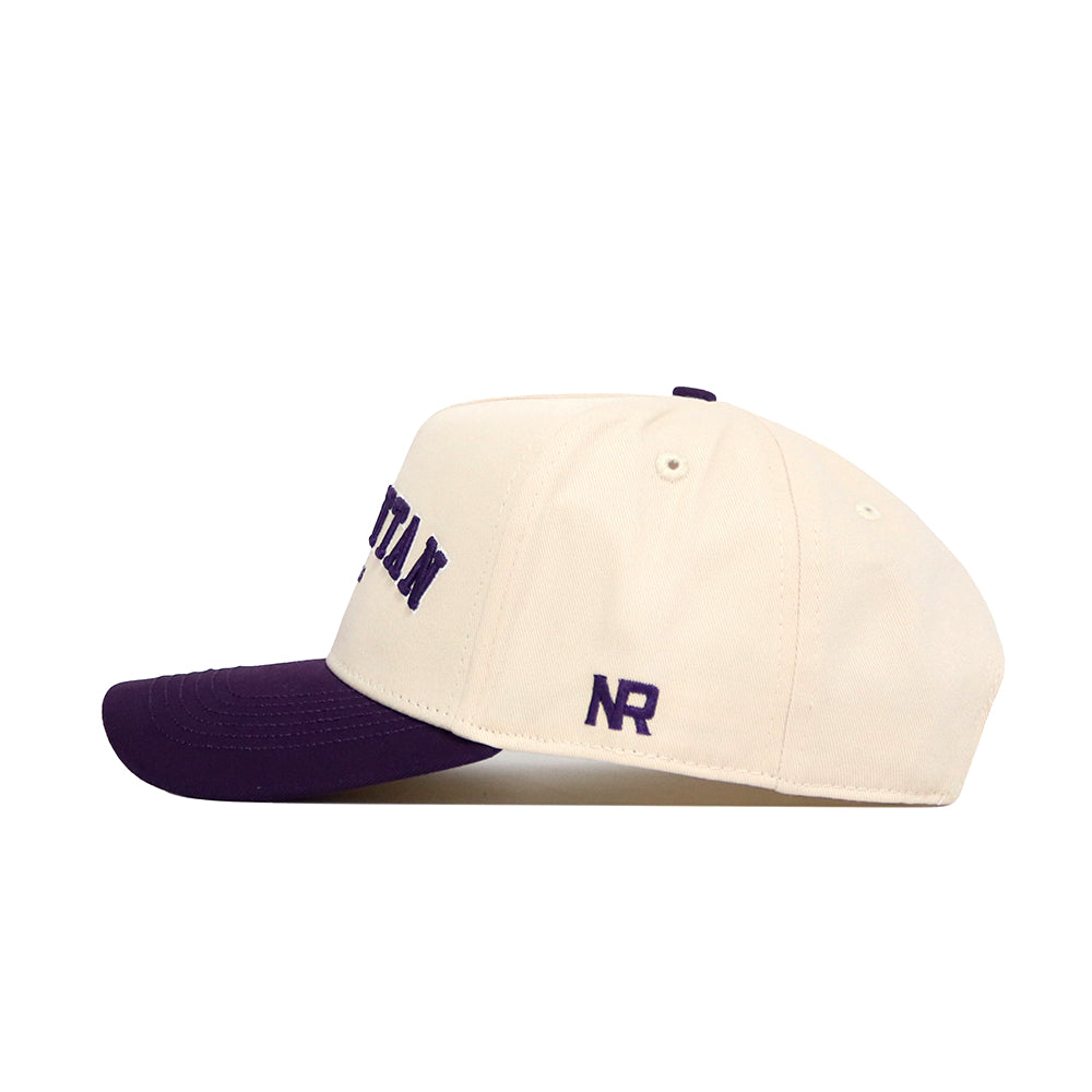 The Manhattan Hometown Hat by No Rivals features a cream structured fit with a dark purple brim, purple NR embroidery on the side, and partial purple text on the front. Made from premium cotton, it's shown in side profile against a white background.