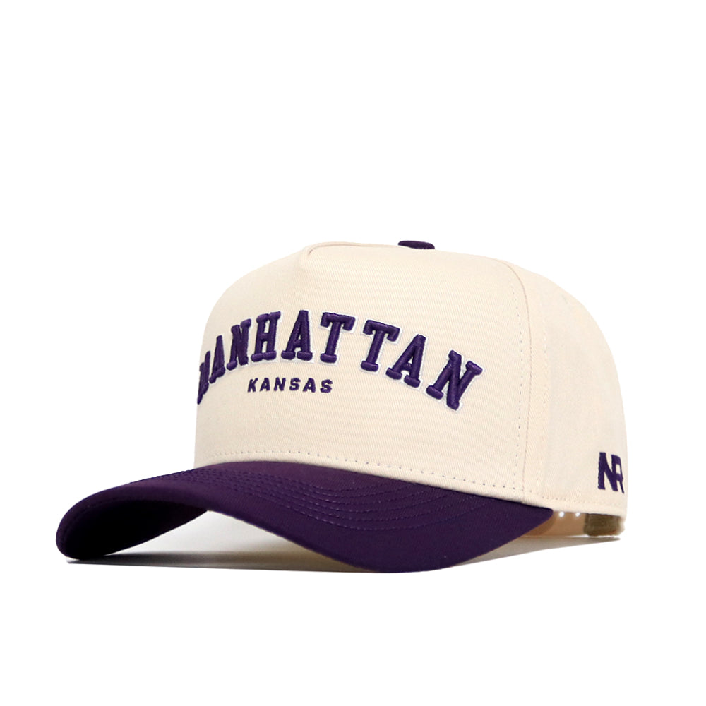 The Manhattan Hometown Hat by No Rivals is a structured beige cotton cap with a purple curved brim, "MANHATTAN KANSAS" in purple and black embroidery, "AR" stitched on the side, and an adjustable snap back for comfort.