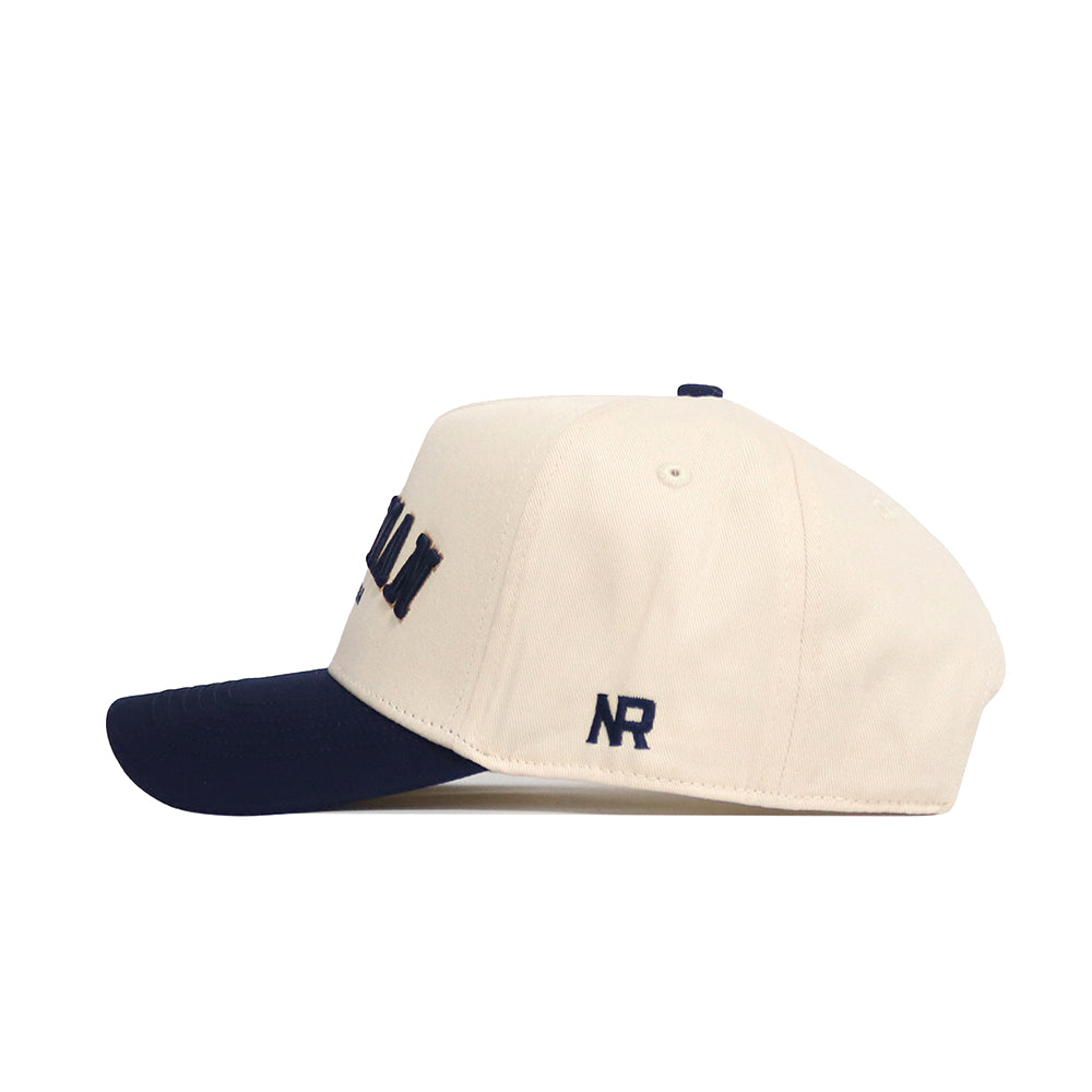 The Bozeman Hometown Hat by No Rivals is a cream cotton baseball cap with a navy brim, embroidered NR letters on the side, ventilation eyelets, structured crown, and an adjustable snap back for comfort.