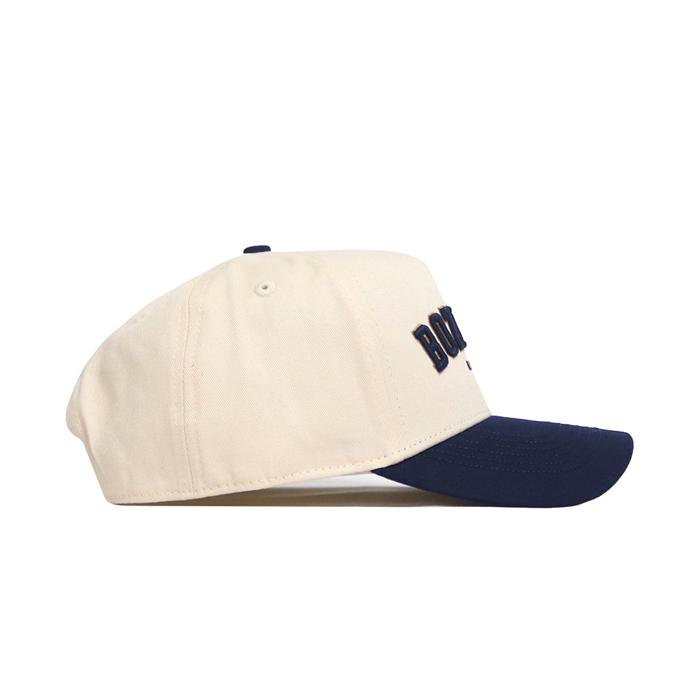 The No Rivals Bozeman Hometown Hat is a beige cotton baseball cap with a navy curved brim, raised front embroidery, and structured fit, shown from the side on a white background.