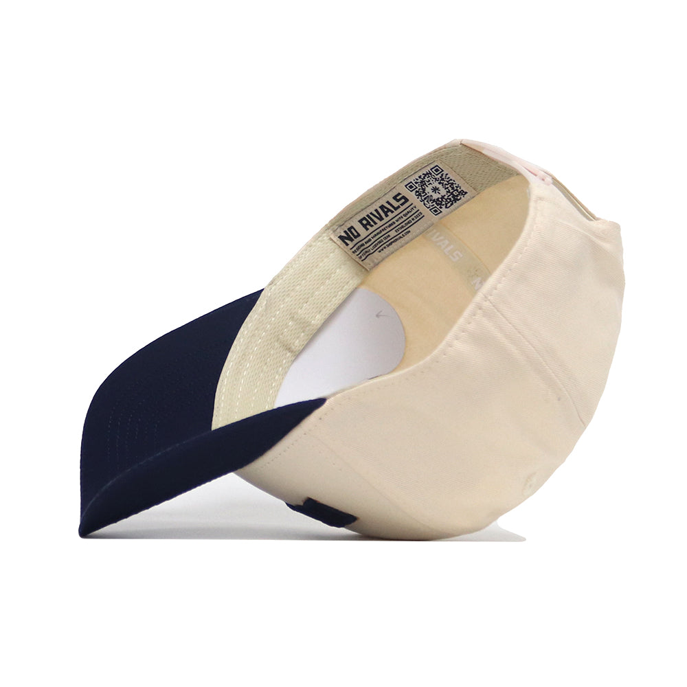 The Bozeman Hometown Hat by No Rivals is shown from the underside, revealing a structured beige cotton cap with a navy blue brim, inner lining, label tag reading NO RIVALS, and a QR code by the band against a white background.