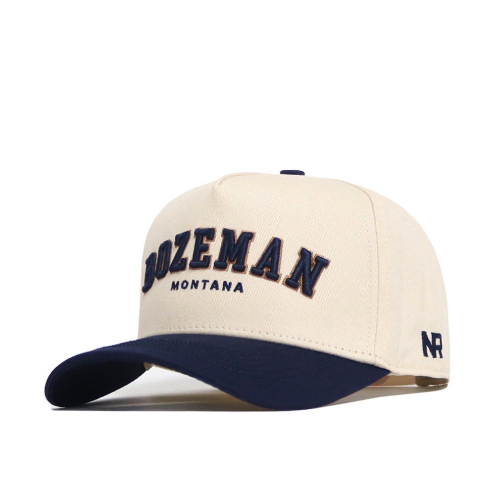 The No Rivals Bozeman Hometown Hat is a cream cap with a navy brim, structured fit, soft cotton, "BOZEMAN MONTANA" embroidered in navy and gold, "NR" on the left, and an adjustable snapback closure.
