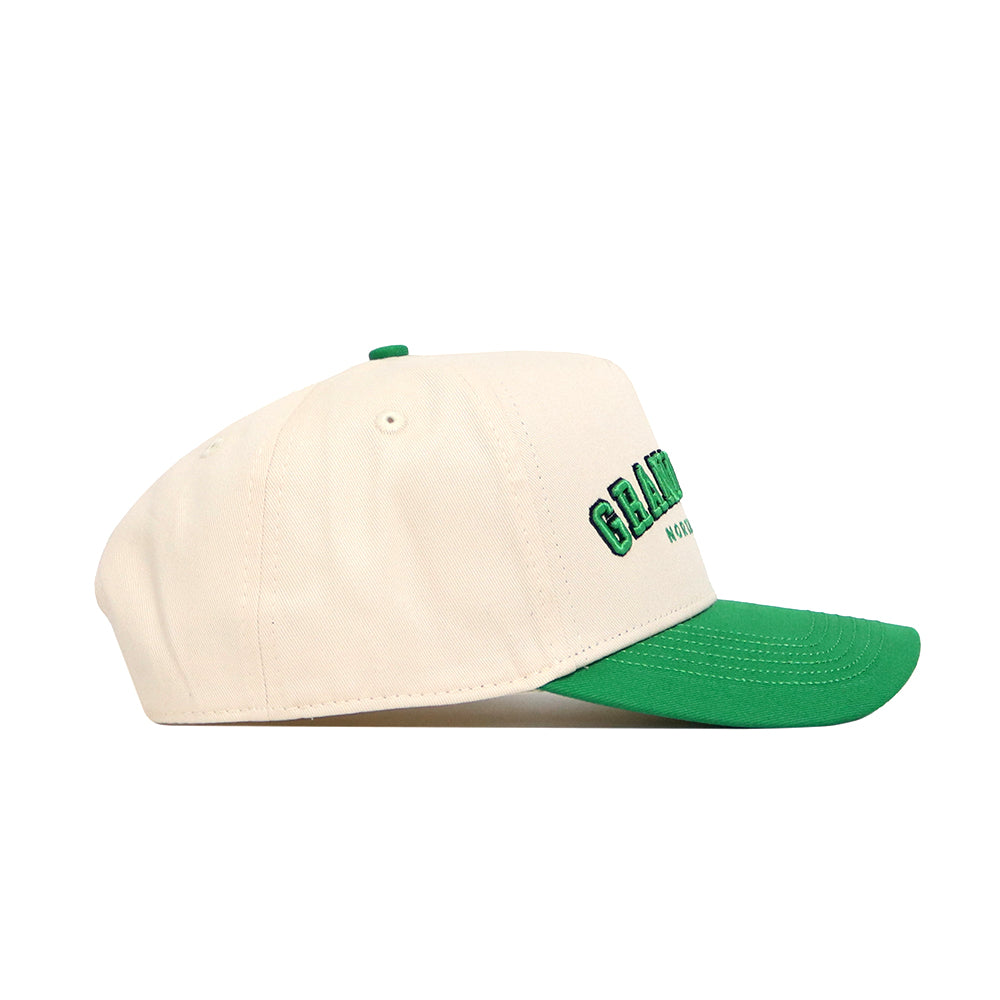 The No Rivals Grand Forks Hometown Hat is a cream snapback with a green brim and button, made of cotton. It features green and black embroidered front text and a structured fit, shown in profile on a white background.