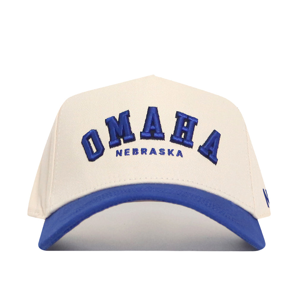 The Omaha Hometown Hat by No Rivals is a white cotton baseball cap with a blue brim, featuring "OMAHA" in large blue letters and "NEBRASKA" in smaller blue letters embroidered on the front.