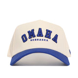 The Omaha Hometown Hat by No Rivals is a white cotton baseball cap with a blue brim, featuring "OMAHA" in large blue letters and "NEBRASKA" in smaller blue letters embroidered on the front.