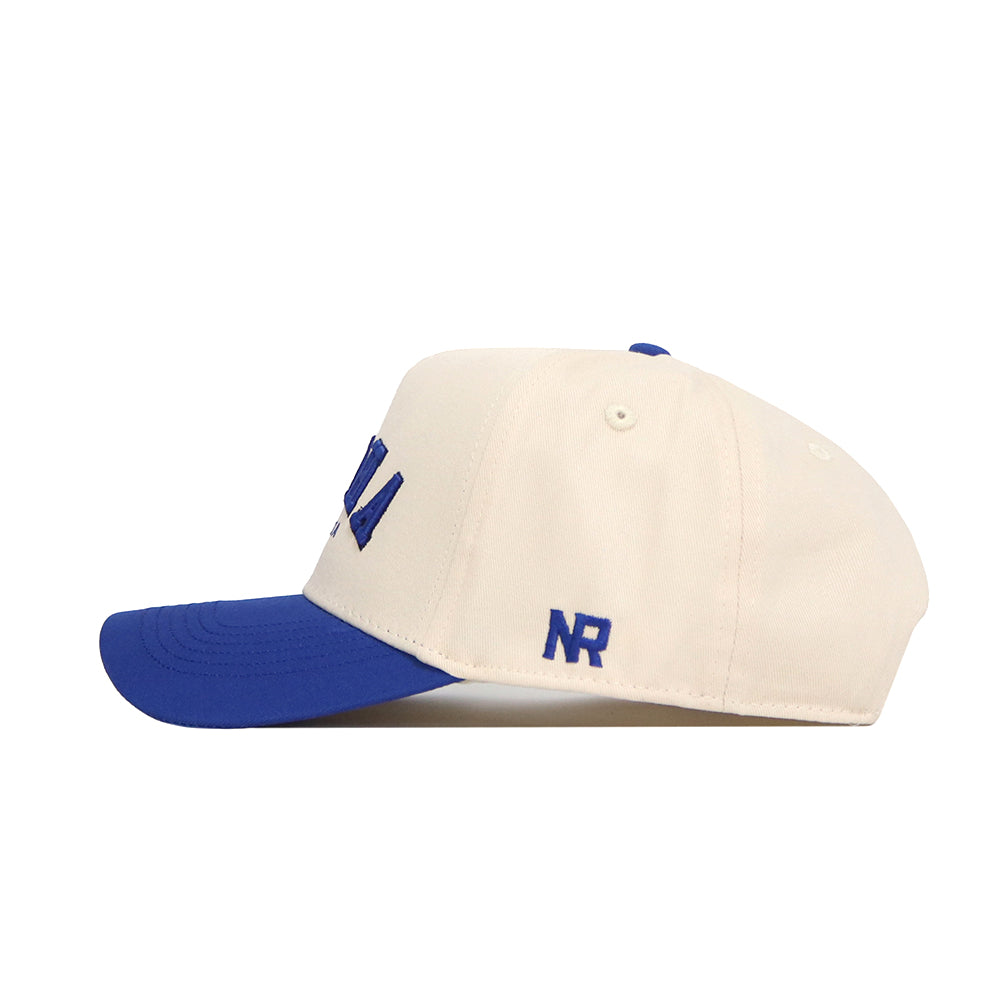 Side view of the Omaha Hometown Hat by No Rivals, a cream-colored structured cotton cap with a blue brim. Blue embroidery accents the front and “NR” is stitched on the side, showcasing classic baseball styling.