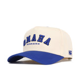 The Omaha Hometown Hat by No Rivals is a cream structured cotton hat with a blue brim, featuring bold blue "OMAHA" and "NEBRASKA" on the front and embroidered NR initials on the side.