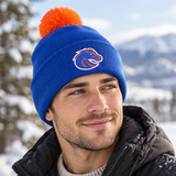 A man wearing the No Rivals Boise State Broncos Logo Beanie, featuring a blue knit with an orange pom-pom and an officially licensed, embroidered horse logo, smiles outdoors in a snowy, mountainous landscape.