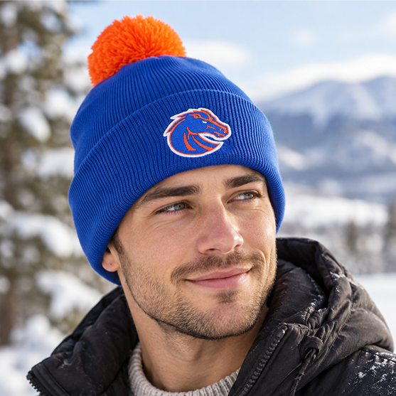 A man wearing the No Rivals Boise State Broncos Logo Beanie, featuring a blue knit with an orange pom-pom and an officially licensed, embroidered horse logo, smiles outdoors in a snowy, mountainous landscape.