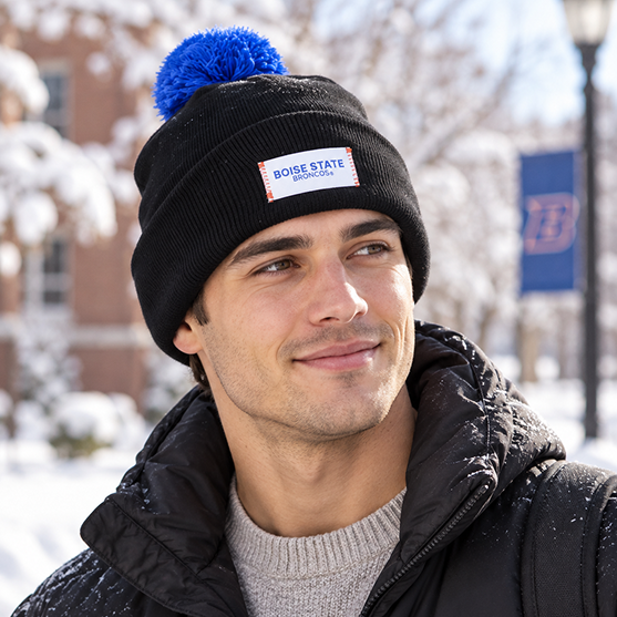 A person stands outside on a snowy winter day, smiling in the No Rivals Boise State Broncos Patch Beanie—an officially licensed black beanie featuring a blue pom-pom and an eye-catching embroidered Broncos logo—creating the perfect gameday look with trees in the background.