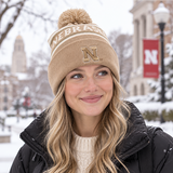 A smiling woman wearing a No Rivals Nebraska Cornhuskers Desert Beanie, featuring an embroidered logo, and a black jacket stands outside in snowy weather with campus buildings and red Nebraska banners in the background.