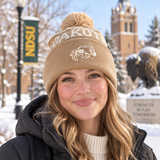 A smiling person wearing the No Rivals North Dakota State Bison Desert Beanie stands outside on a snowy day with a bison statue, trees, and an NDSU banner in the background, proudly showing off their officially licensed gameday fit.