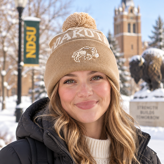 A smiling person wearing the No Rivals North Dakota State Bison Desert Beanie stands outside on a snowy day with a bison statue, trees, and an NDSU banner in the background, proudly showing off their officially licensed gameday fit.
