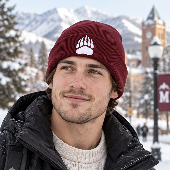 A young man wears the No Rivals Montana Grizzlies Logo Beanie, featuring an officially licensed white bear paw emblem, as he smiles outdoors in a snowy scene with mountains, trees, and a clock tower in the background.