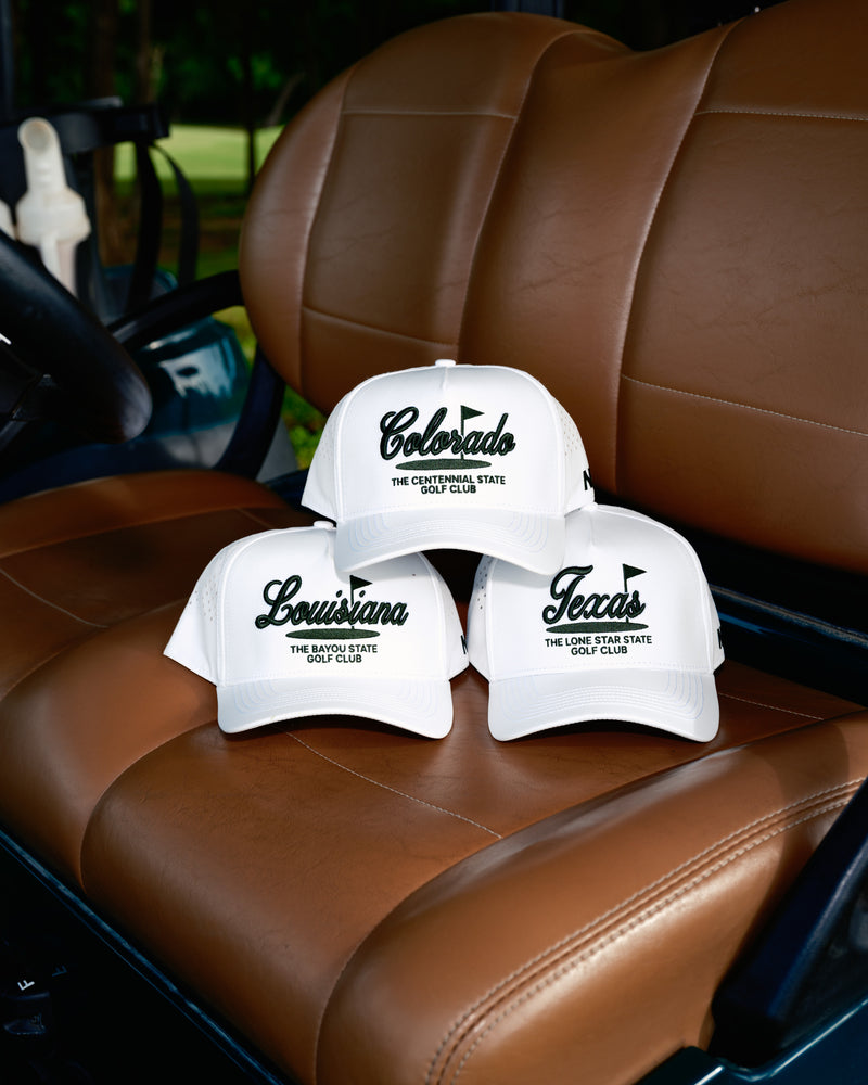 Three white baseball caps with embroidered state names—Colorado, Louisiana, and Texas—rest on a brown leather seat of a golf cart, outdoors with greenery in the background.