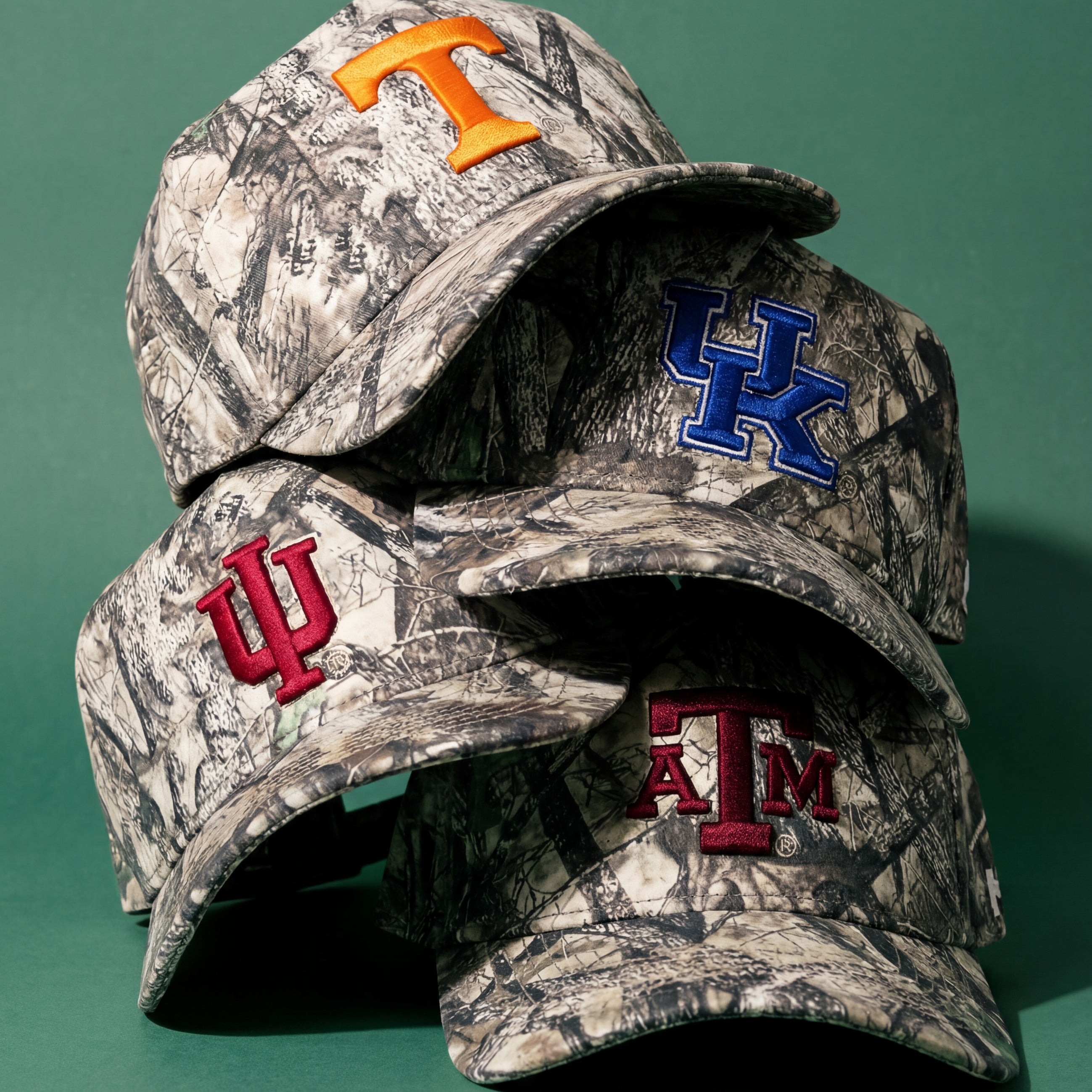 Four camouflage-patterned baseball caps featuring embroidered college logos: Tennessee (orange T), Kentucky (blue UK), Indiana (red IU), and Texas A&M (maroon ATM), stacked against a green background.