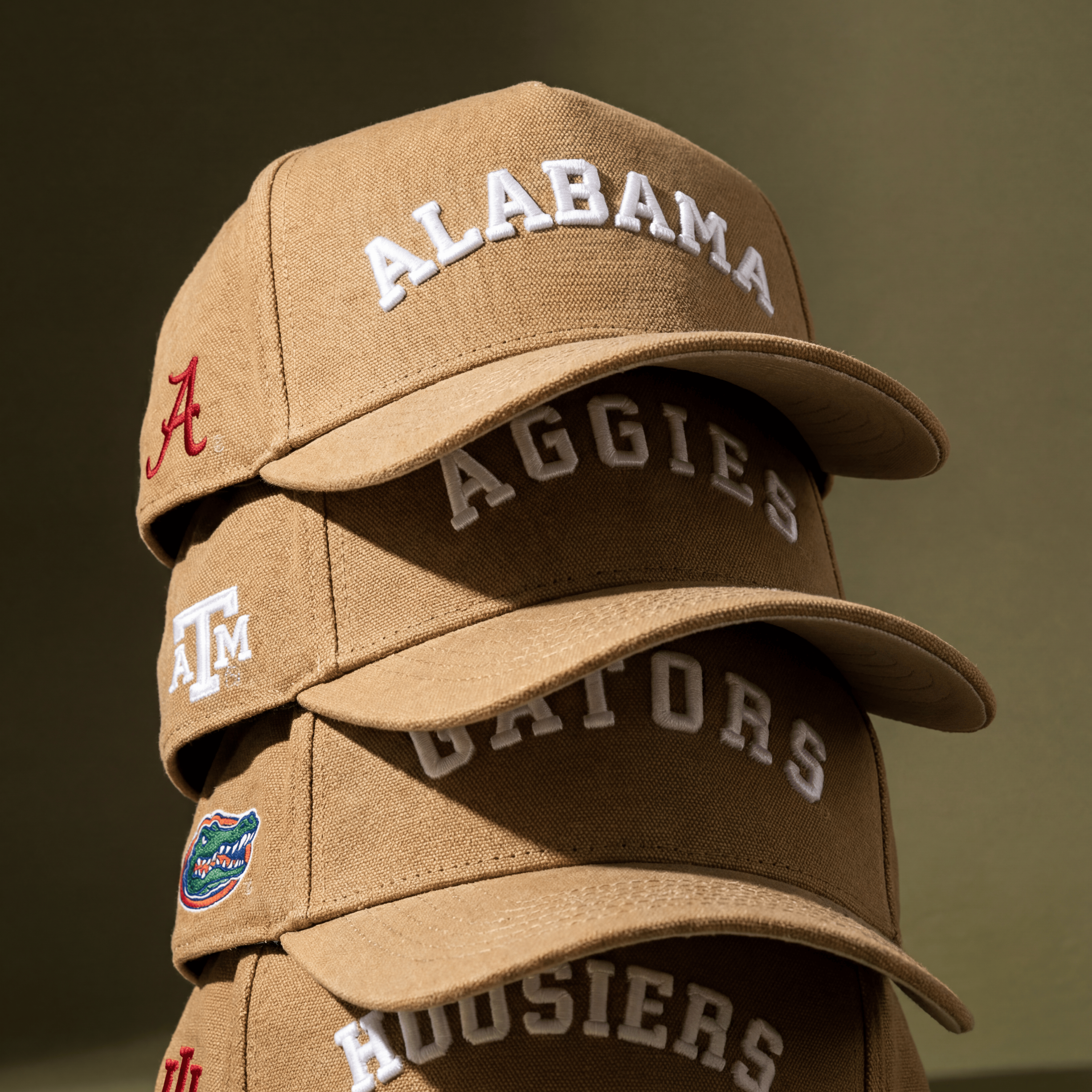 Four brown baseball caps stacked vertically, each embroidered with a different college name: Alabama, Aggies, Gators, and Hoosiers. The caps have logos and text in white and red on a greenish-brown background.