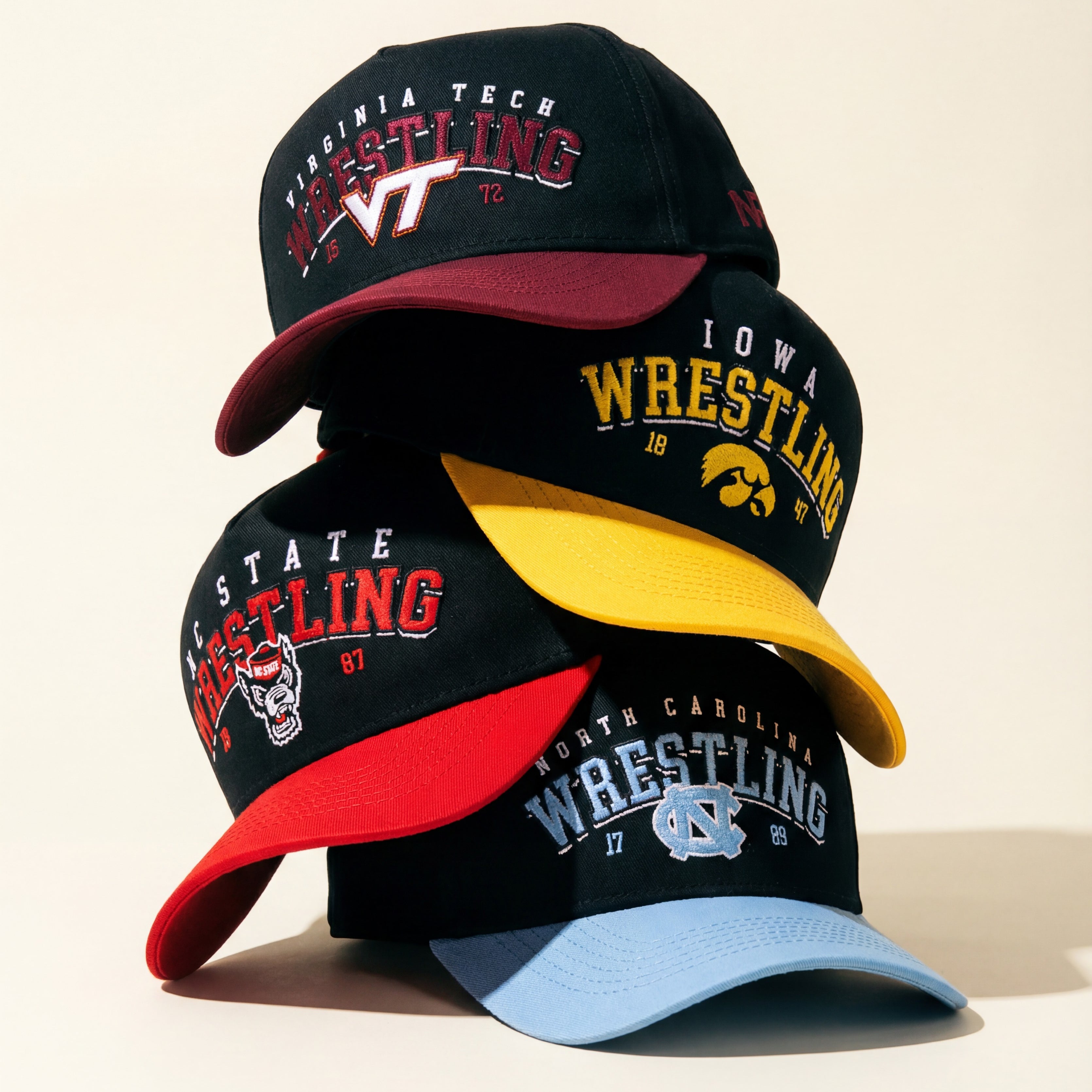 Four wrestling caps stacked. Top cap is black for Virginia Tech, second is black and yellow for Iowa, third is black and red for NC State, and bottom is navy and light blue for North Carolina. All have embroidered logos.