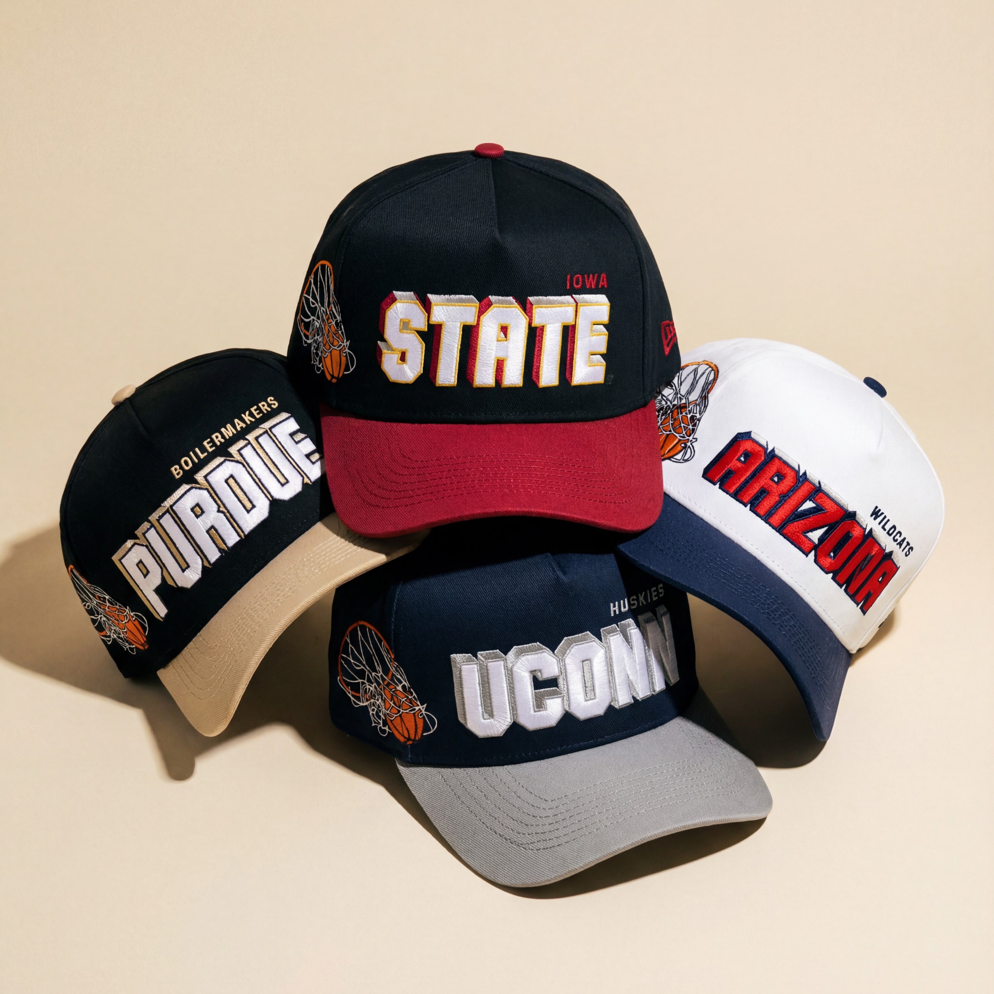 Four embroidered baseball caps feature the names Purdue, State, UConn, and Arizona, arranged on a beige surface. Each cap has unique team colors and a basketball graphic on the side.