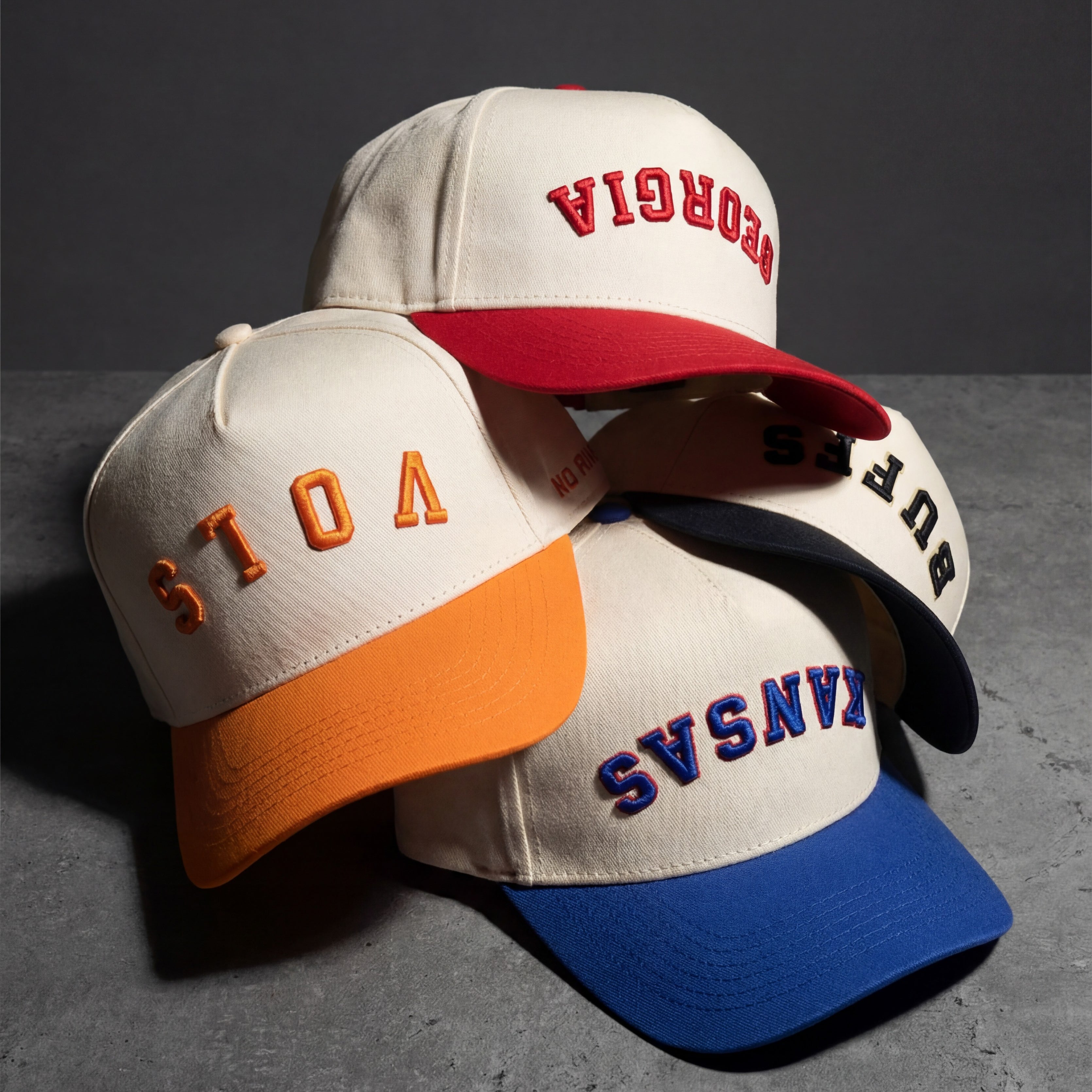 Four baseball caps stacked on a gray surface. The caps are cream-colored with colored brims and embroidered words: VOTE (orange), GEORGIA (red), BURBS (black), and KANSAS (blue).