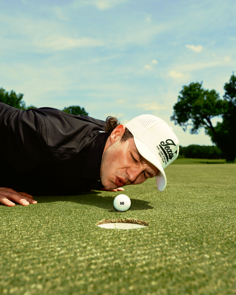 A man in a black shirt and white cap is lying on the grass of a golf course, closely eyeing a golf ball near the hole with his cheek almost touching the ground under a blue sky.