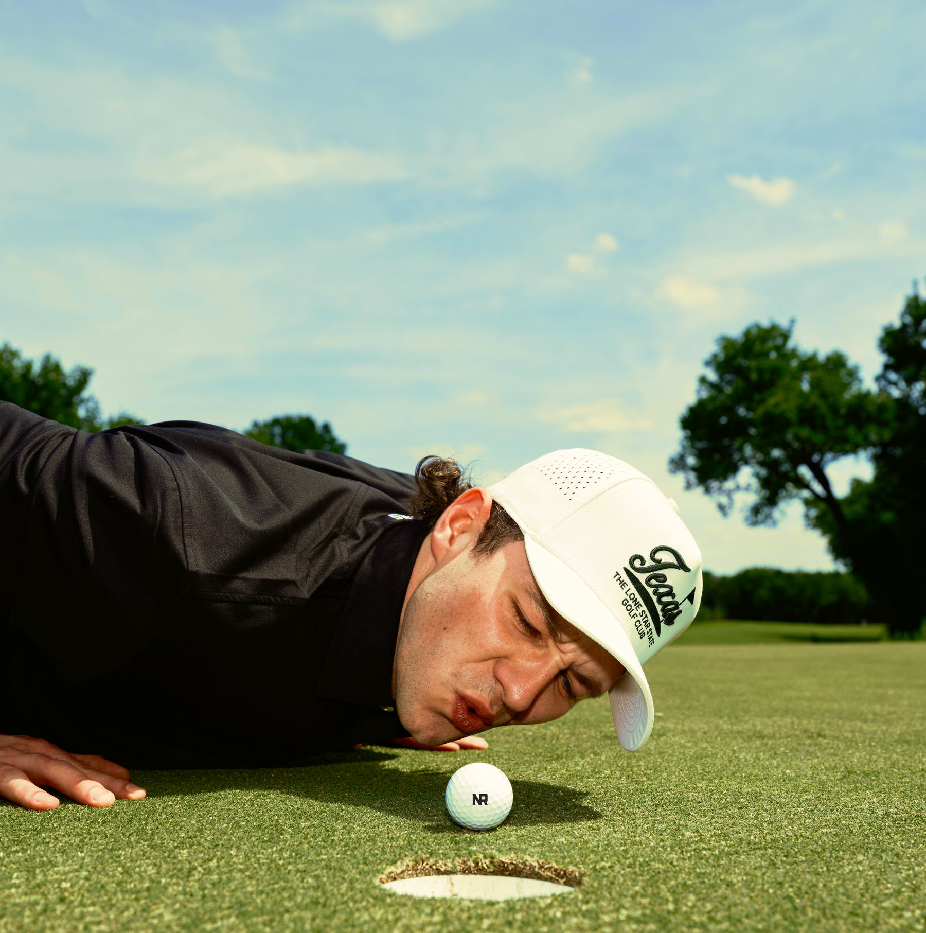 A man in a black shirt and white cap is lying on the grass of a golf course, closely eyeing a golf ball near the hole with his cheek almost touching the ground under a blue sky.
