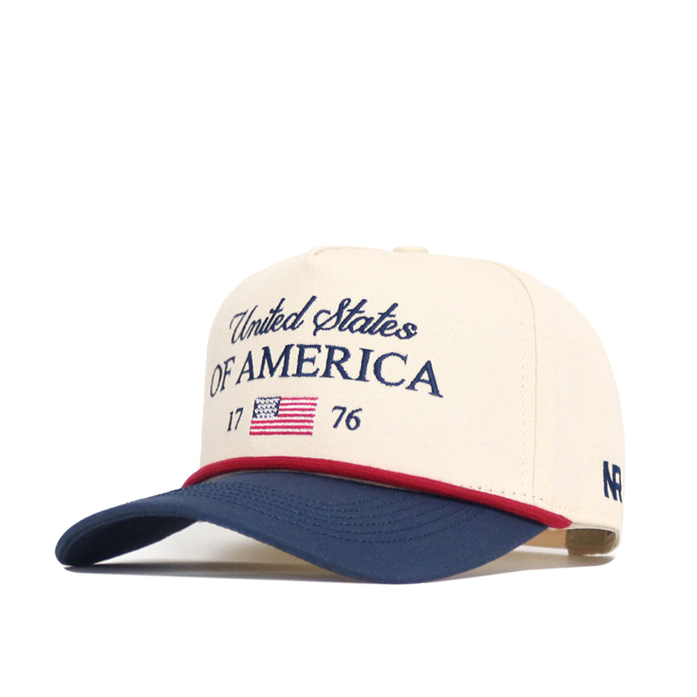 The No Rivals USA Classic Americana Hat features cream cotton, "United States of America 1776" in navy embroidery, a small American flag patch, a navy curved brim with red trim, and offers a structured snap back fit.
