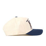 A side view of the No Rivals WVU Mountaineers Stacked Mascot Hat, a cream-colored baseball cap with a navy blue brim and an embroidered jumping mascot logo on the side panel.