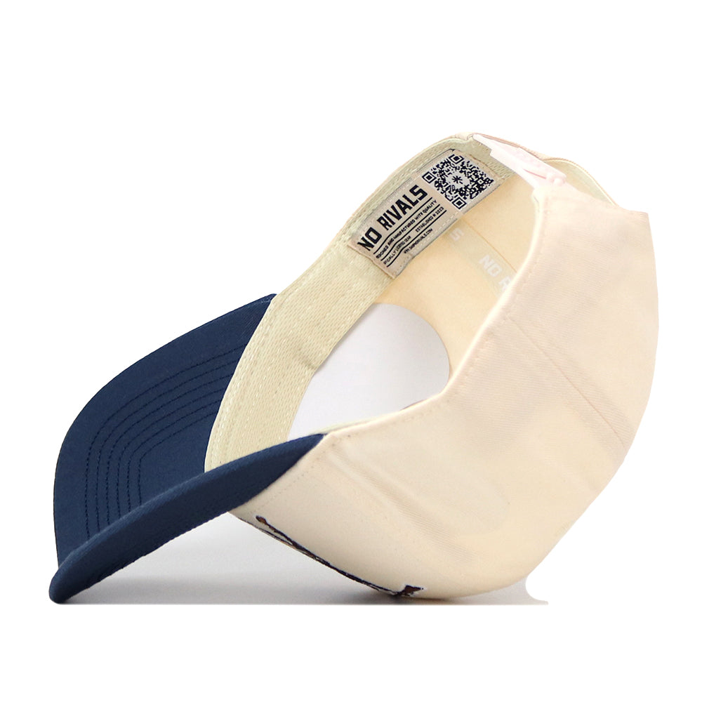 A WVU Mountaineers Stacked Mascot Hat by No Rivals in cream and navy blue is shown upside down, highlighting its inner label, QR code, and adjustable strap against a white background. The hat is crafted from performance fabric for enhanced comfort and durability.