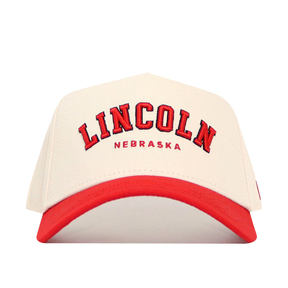 The Lincoln Hometown Hat by No Rivals is a cream structured cotton cap with a red brim, bold LINCOLN NEBRASKA embroidery on the front, and an adjustable snap back for a custom fit.