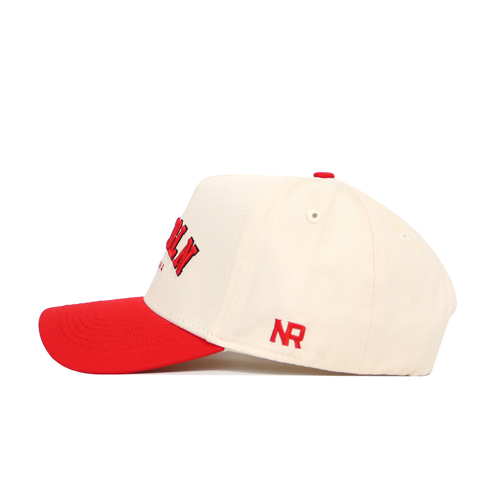 The Lincoln Hometown Hat by No Rivals is a cream structured fit cap crafted from high quality cotton, with a red brim and red embroidered NR logo on the side, shown from a side angle.