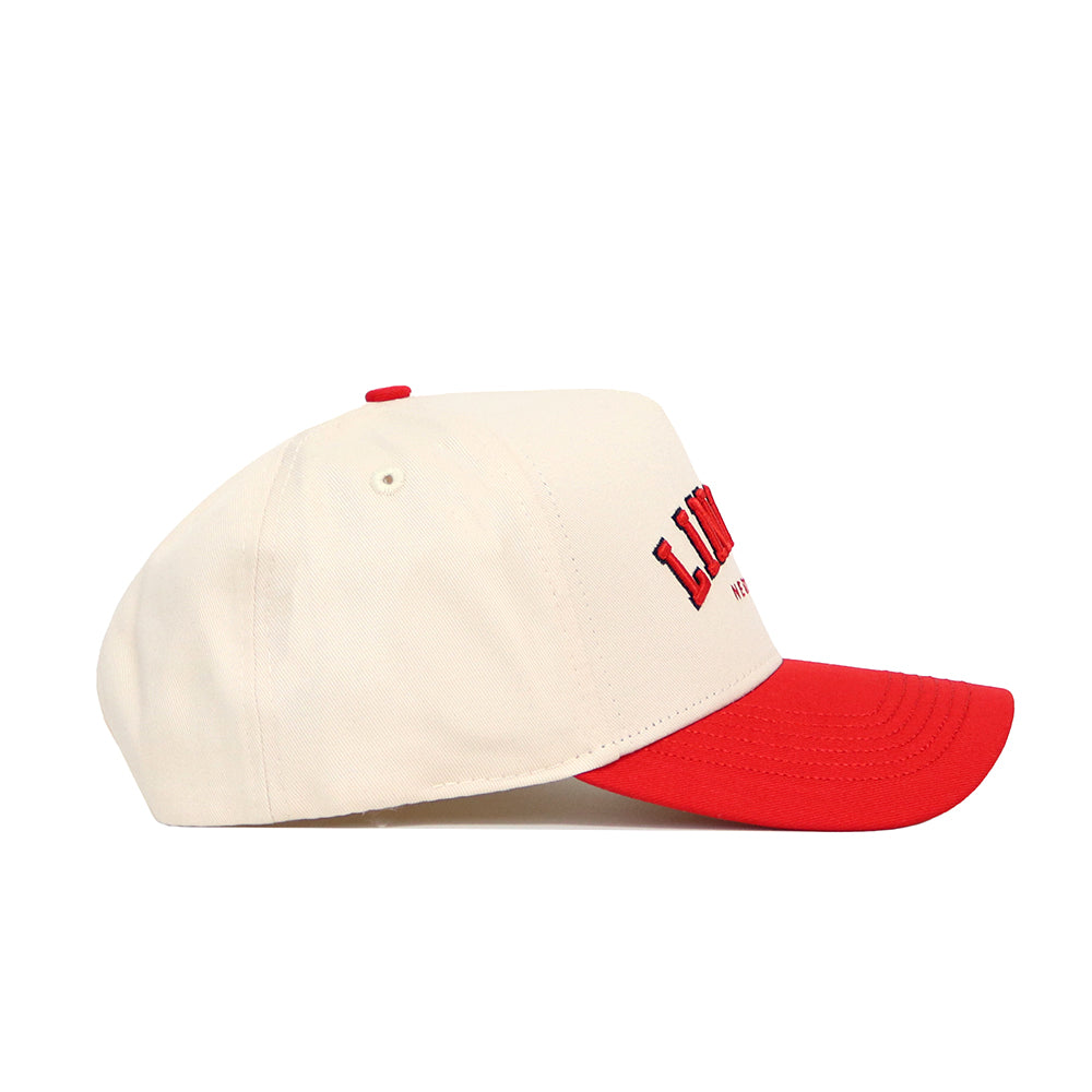 The Lincoln Hometown Hat by No Rivals is a cream cotton baseball cap with a bright red brim and top button. It has red and black embroidered text on the side. The hat offers a structured fit, shown against a plain white background.