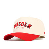 The Lincoln Hometown Hat by No Rivals features a cream snapback design with a red brim and embroidered "LINCOLN NEBRASKA" text, crafted from high-quality cotton for a structured fit.