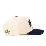 Side view of the No Rivals Georgia Tech Yellow Jackets Stadium Hat, featuring a beige crown with visible stitching and ventilation holes and a dark navy brim. A gold and black GT logo is embroidered on the side. The snap back adjustment ensures a perfect gameday fit.