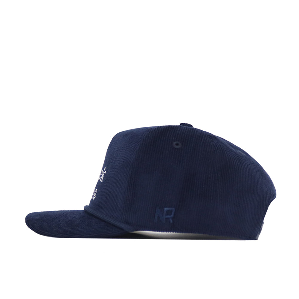 Side view of the No Rivals Back Nine Cord Hat in navy. This cotton corduroy baseball cap features a flat brim, subtle embroidered logos near the back, a snapback closure, and a button on top. Background is plain white.