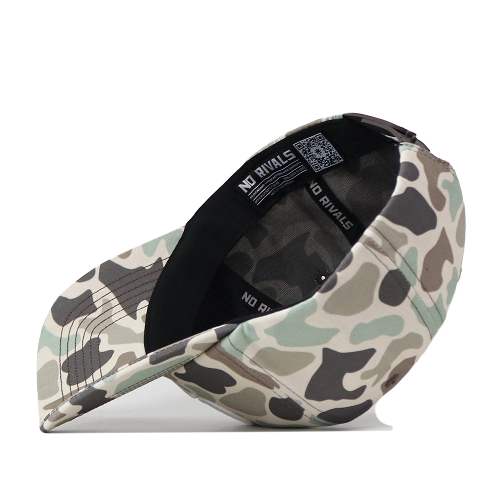 A Montana State Script Huntsman Hat by No Rivals is shown upside down, displaying the inner lining with NO RIVALS text and a QR code on the inside band. This snapback cap features a classic camouflage pattern for a bold, rugged look.