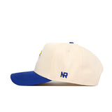 The No Rivals Kansas Logo Patch Hat is a cream cotton cap with a blue brim, blue top button, NR initials embroidered on the side, detailed front patch embroidery, and a classic snapback closure.