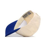 The Kansas Logo Patch Hat by No Rivals is a cream-colored cotton blend snapback with a blue brim, shown inside out to display a patch reading NO RIVALS and a QR code sewn into the inner band.
