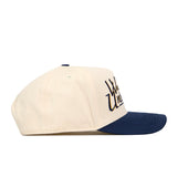A side view of the No Rivals WVU Mountaineers Script Retro Hat showcases its cream-colored crown and navy blue brim. The front panel features a partially visible dark embroidered logo, while the classic snapback closure offers an adjustable fit.