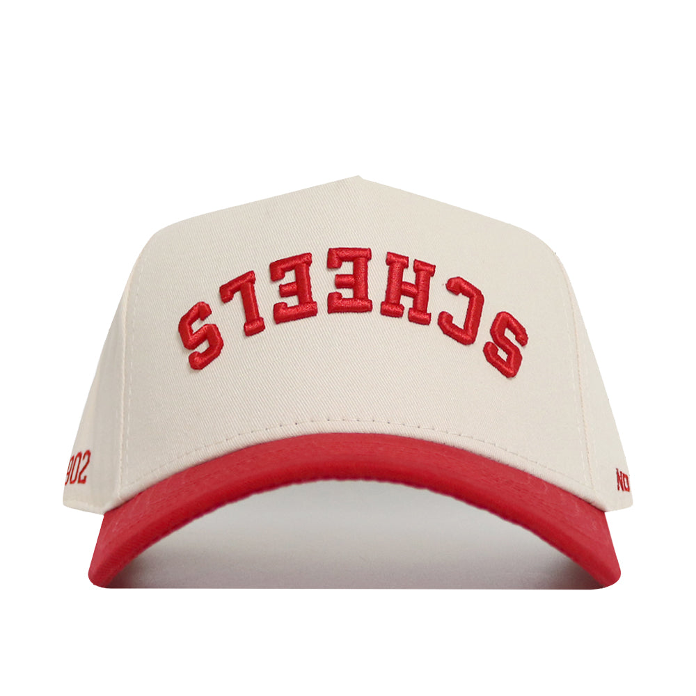 The Upside Down Scheels Hat is a white cotton baseball cap with a red brim, red upside-down embroidered SCHEELS lettering on the front, and an adjustable snap back closure.