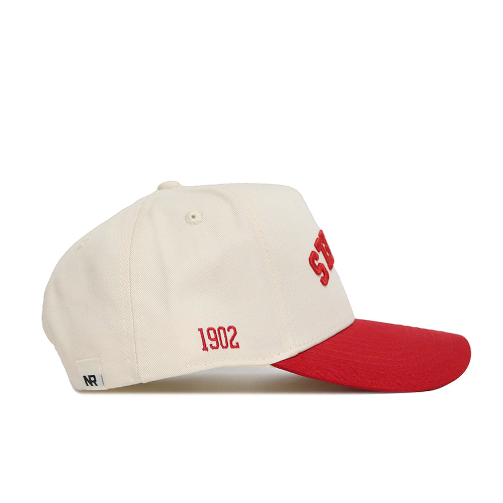 The Upside Down Scheels Hat is a cream baseball cap with a red brim, made from high-quality cotton. It features "1902" embroidered in red on the side, partial red lettering on the front, a small white tag at the back, and an adjustable snap closure.